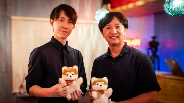 Two men in black shirts smile while holding light brown teddy bears with red ribbons, indoors at a colorful venue.