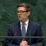 Man in a dark suit and glasses speaks at a lectern against a green marble backdrop, delivering a formal speech.