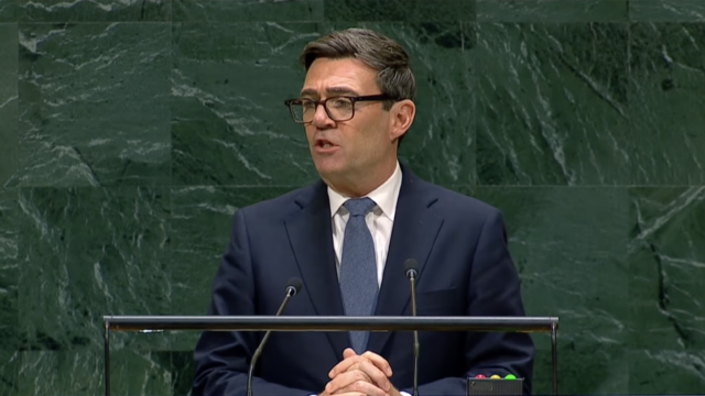 Man in a dark suit and glasses speaks at a lectern against a green marble backdrop, delivering a formal speech.