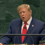Man in a dark suit and red tie speaks at a podium with microphones against a green marble backdrop.