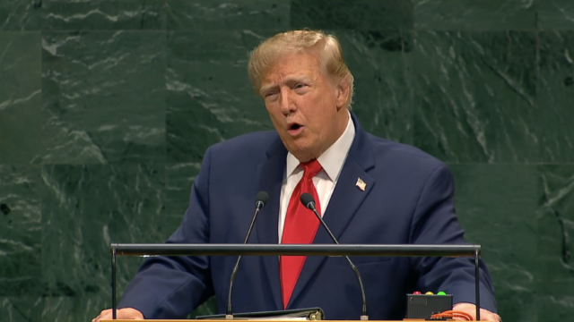 Man in a dark suit and red tie speaks at a podium with microphones against a green marble backdrop.