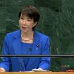 Woman in a blue suit delivering a speech at a wooden podium with microphones in front of a green marble backdrop.