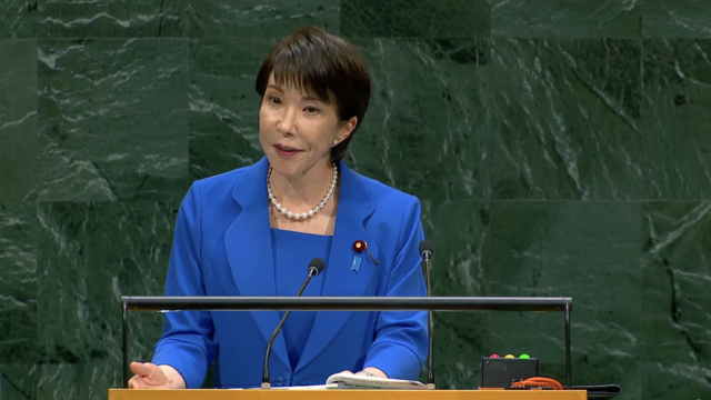 Woman in a blue suit delivering a speech at a wooden podium with microphones in front of a green marble backdrop.