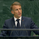 Man in a dark suit speaks at a podium with microphones, UN emblem visible in the top right corner behind him.
