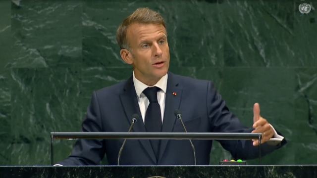 Man in a dark suit speaks at a podium with microphones, UN emblem visible in the top right corner behind him.