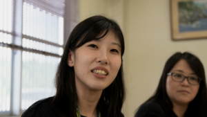 Close-up of a smiling young woman with dark hair, with a second person partially visible on the right indoors.